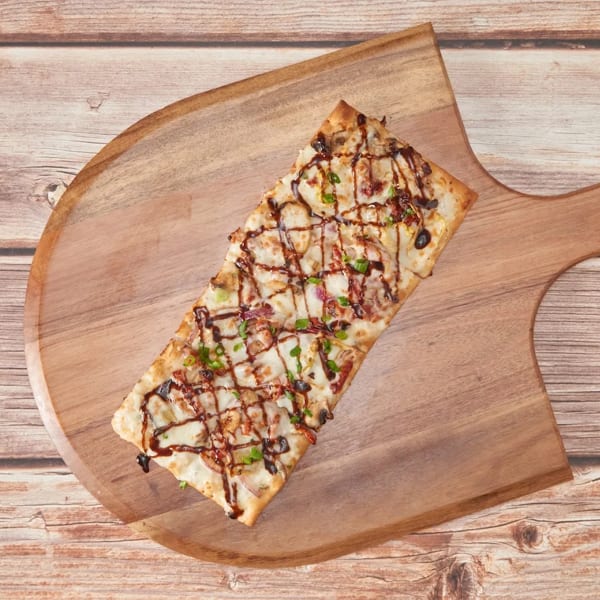 Rectangular flatbread pizza with white sauce, cheese, bacon, green onions, and a balsamic glaze drizzle on a wooden board.
