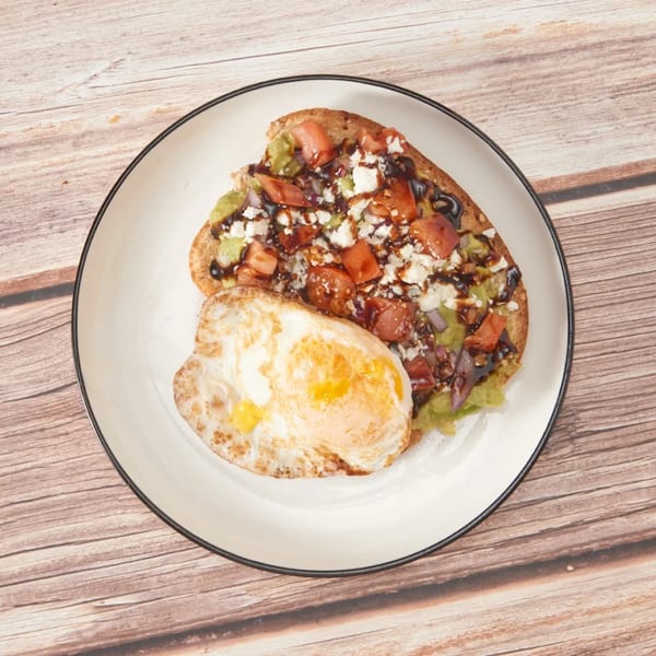 Avocado toast with tomatoes, feta, balsamic glaze, and a fried egg on a white plate.