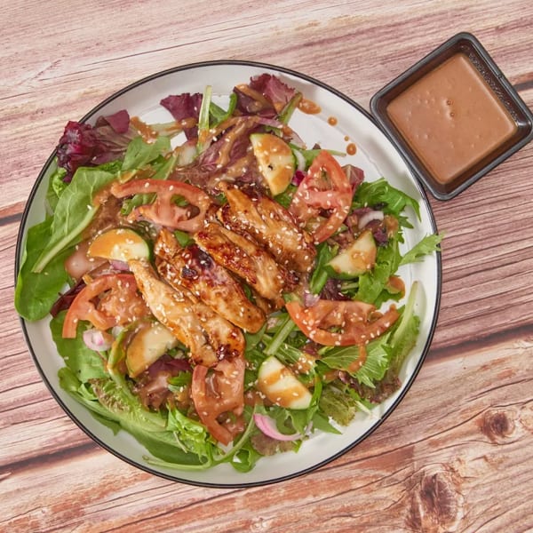 Teriyaki chicken salad with mixed greens, tomatoes, cucumbers, and sesame seeds, with a side of dressing.